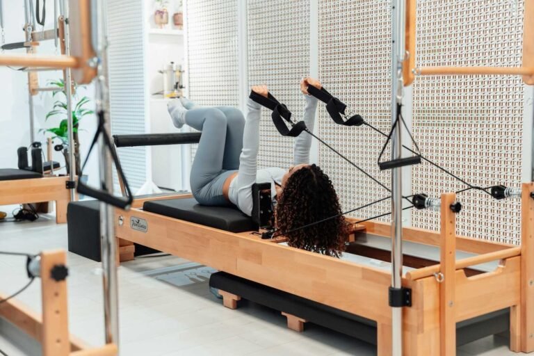 Woman practicing supine Pilates on a reformer using arm straps in a bright Pilates studio.