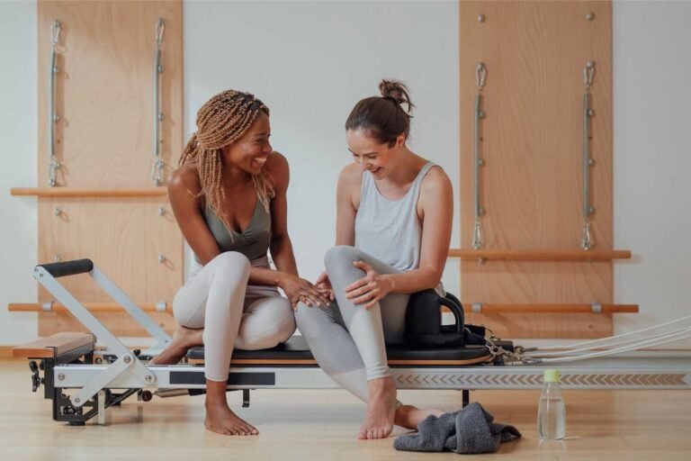 Two women smiling on a Pilates reformer practicing affirmations for body image in a bright studio.